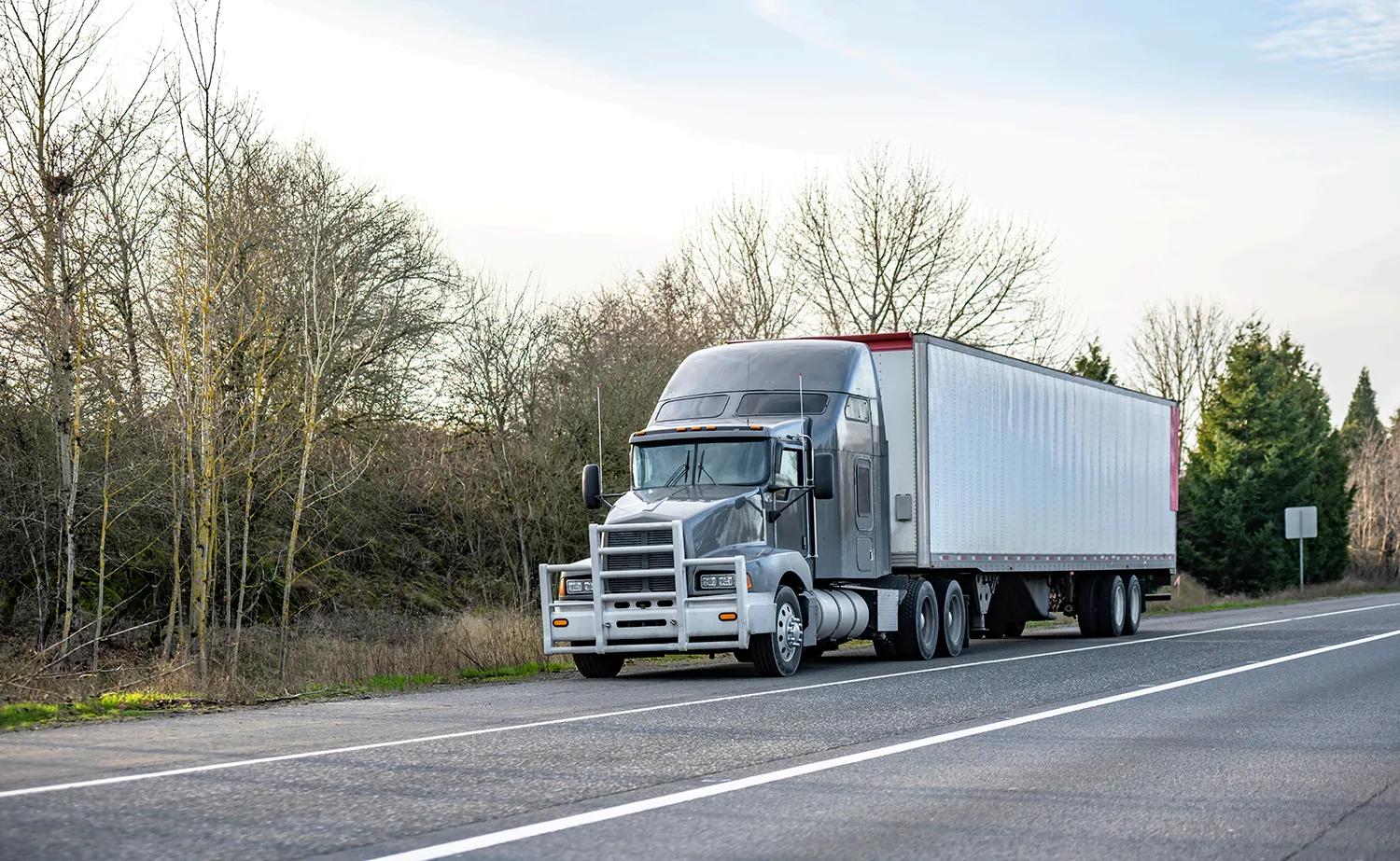 Gray semi-truck with a large silver trailer parked on the roadside beside a two-lane highway, leafless trees in the background.