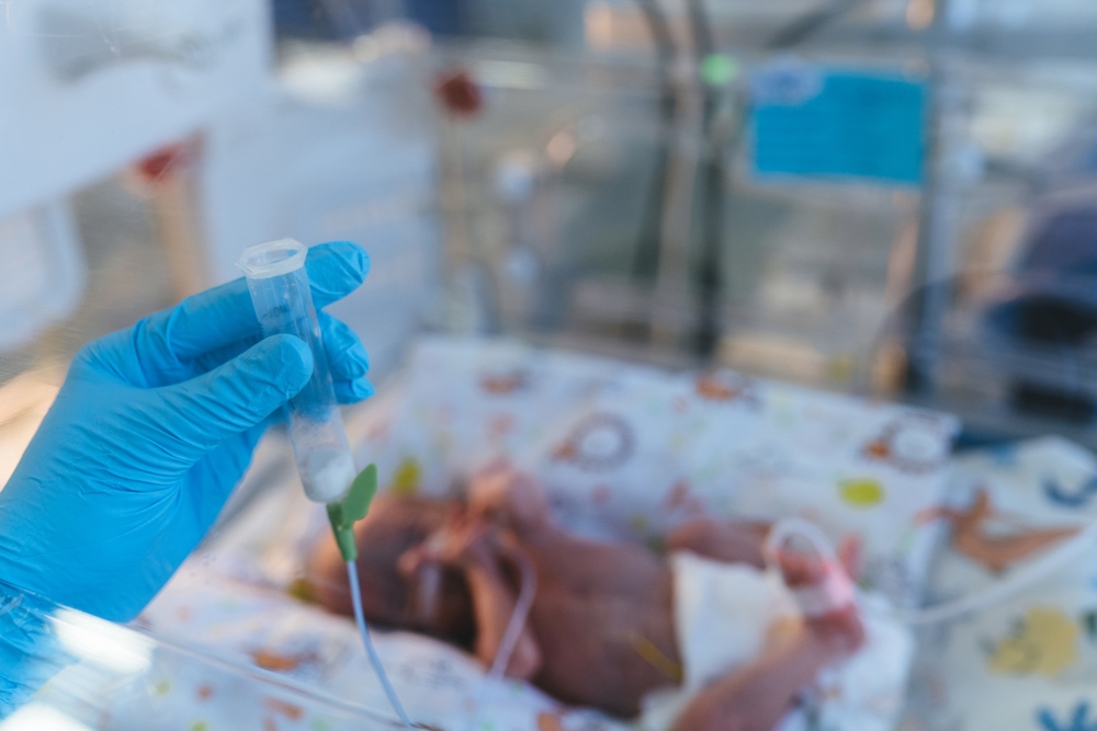 Gloved hand holding a syringe beside a newborn in an incubator in a hospital setting