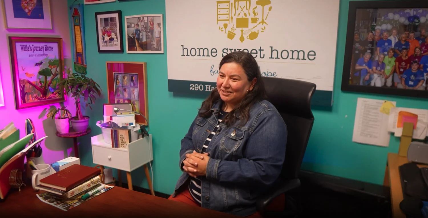 Woman seated at a desk in a colorful office, hands folded, smiling with a large 'home sweet home' sign on the teal wall behind her.